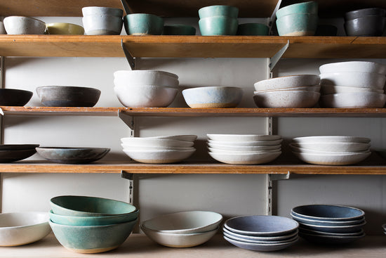 Stacks of sage, ivory, black, and sapphire bowls sit on rustic wooden shelves
