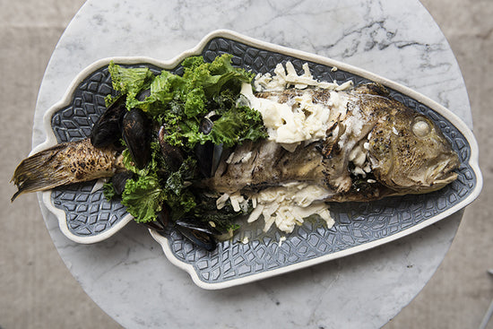 A roasted branzino garnished with greens and shaved cheese rests on a textured, fish-shaped platter